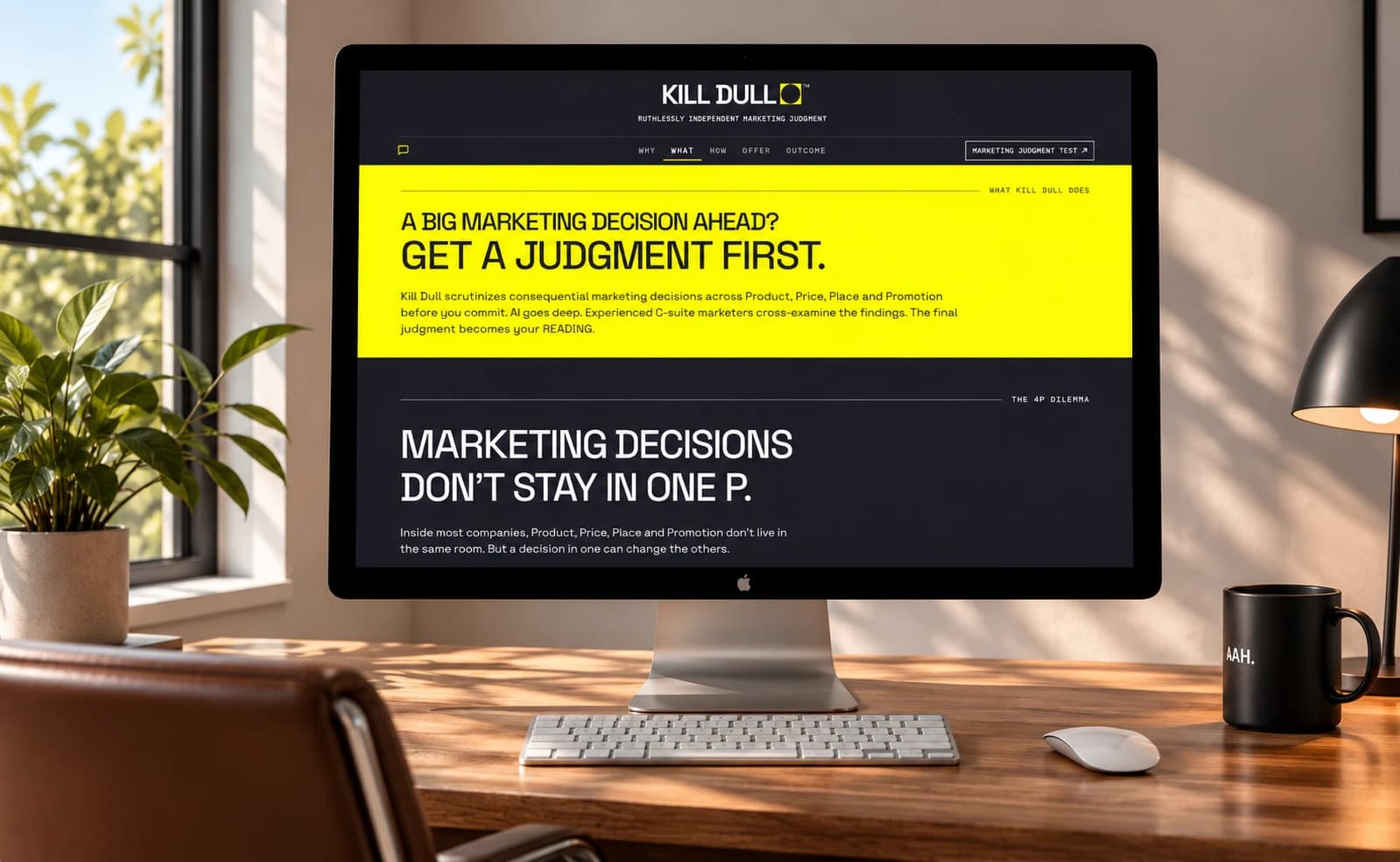 The Kill Dull website on a desktop monitor, on a sunlit wooden desk beside a plant and an AAH. mug